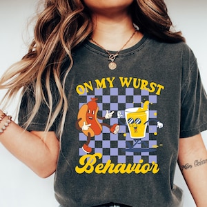 May include: A gray t-shirt with a graphic of a cartoon hot dog and a glass of beer on a checkered background. The text "On My Wurst Behavior" is printed on the shirt.