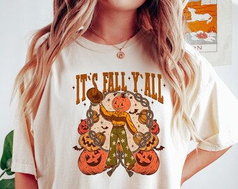 May include: Cream-colored t-shirt with the text "IT'S FALL Y'ALL" in brown and orange. The graphic features a pumpkin-headed scarecrow holding a hat, surrounded by pumpkins and chains. The shirt is a relaxed fit.