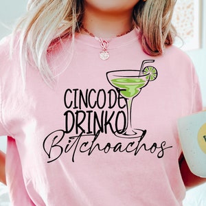 以下が含まれることがあります： ピンクのTシャツに「Cinco de Drinko Bitchoachos」の文字と、ライムのくさび形がついた緑色のマルガリータグラスがプリントされています。