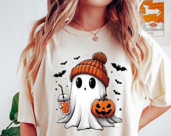 May include: A white t-shirt with a cartoon ghost wearing an orange and brown knit hat, holding a pumpkin and a cup of coffee. The ghost is surrounded by bats and stars.