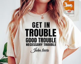 May include: A cream-coloured t-shirt featuring the text "GET IN TROUBLE GOOD TROUBLE NECESSARY TROUBLE" and the signature "John Lewis" in black. The shirt is paired with dark green shorts. A bracelet is visible on the left wrist.