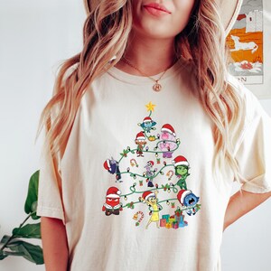 May include: A white t-shirt with a Christmas tree design featuring characters from the movie Inside Out. The tree is decorated with lights, ornaments, and candy canes. The characters are wearing Santa hats and are arranged in a festive tree shape.