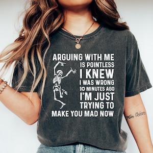May include: Dark gray t-shirt with the text "ARGUING WITH ME IS POINTLESS I KNEW I WAS WRONG 10 MINUTES AGO I'M JUST TRYING TO MAKE YOU MAD NOW" and a skeleton graphic. The shirt is paired with blue jeans and white shoes.