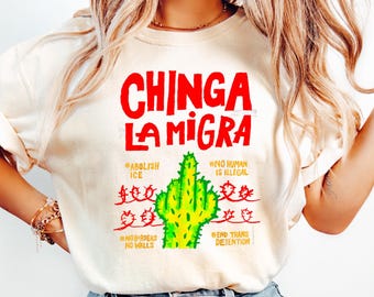 May include: Cream-colored t-shirt with the red text "CHINGA LA MIGRA" above a green cactus illustration. Additional red text includes "#ABOLISH ICE", "#NO HUMAN IS ILLEGAL", "#NO BORDERS NO WALLS", and "#END TRANS DETENTION". The shirt is paired with blue jeans.