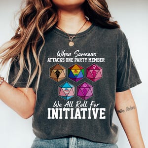 May include: Dark gray t-shirt with the text "When Someone Attacks One Party Member, We All Roll For Initiative." The shirt features colorful dice with various symbols. The shirt is worn with blue jeans and white shoes.
