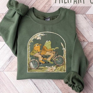 Puede incluir: Una sudadera verde con un gráfico de dos ranas montando una bicicleta. Las ranas llevan ropa y la bicicleta tiene una cesta. El texto "MILITARY GREEN" está impreso en blanco en la parte superior de la imagen.