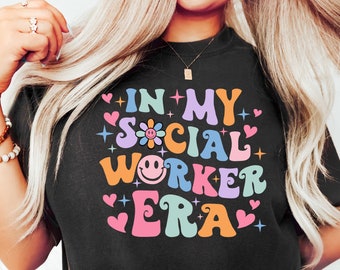 Könnte beinhalten: Schwarzes T-Shirt mit dem Text "IN MY SOCIAL WORKER ERA" in einem farbenfrohen Retro-Schriftzug. Das Design beinhaltet ein Smiley, Herzen und Blumen. Das Shirt ist ein lässiger, bequemer Stil.
