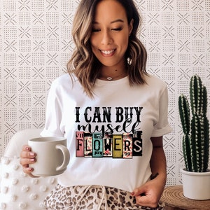 May include: White t-shirt with the text "I can buy myself flowers" in black lettering. The text is surrounded by colorful squares with floral patterns.