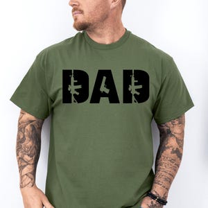 May include: Olive green t-shirt with the word "DAD" in bold black letters. The letters are stylized with gun silhouettes. The shirt is short-sleeved and made of a soft material.