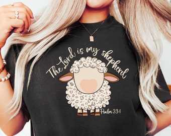 Könnte beinhalten: Schwarzes T-Shirt mit einem weißen und braunen Leopardenmuster-Schaf-Motiv. Der Text "The Lord is my shepherd" ist in goldener Schrift über dem Schaf geschrieben. Der Text "Psalm 23:1" ist in kleinerer goldener Schrift unter dem Schaf geschrieben.