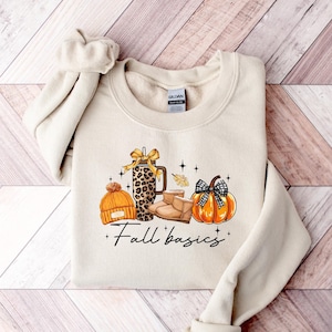 May include: A beige sweatshirt with a graphic of fall-themed items: a pumpkin, boots, a hat, and a leopard-print tumbler. The words "Fall basics" are written in a script font below the graphic. The sweatshirt is folded neatly.
