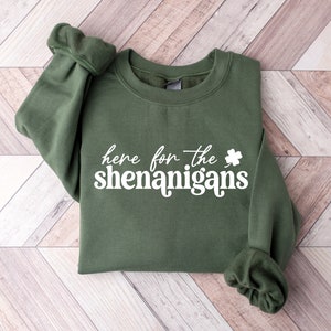 Puede incluir: Una sudadera verde con el texto "here for the shenanigans" y un trébol impreso en blanco.