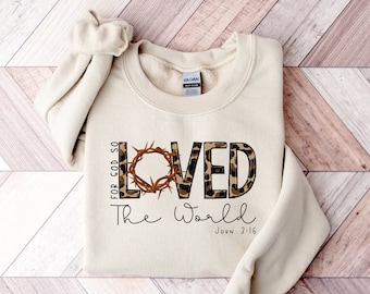 May include: A beige sweatshirt with a leopard print design that says "Loved" with a crown of thorns around the "O". The text "For God so loved the world" is written vertically on the left side of the sweatshirt. The text "John 3:16" is written below the word "world".