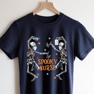 Puede incluir: Una camiseta azul marino con un gr&aacute;fico de dos esqueletos vestidos de enfermeras con mascarillas faciales y sosteniendo suministros m&eacute;dicos. El texto "Spooky Nurse" est&aacute; impreso en naranja en la camiseta.