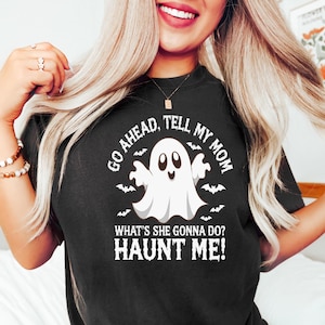 Puede incluir: Camiseta negra con un diseño de fantasma blanco y el texto "GO AHEAD, TELL MY MOM WHAT'S SHE GONNA DO? HAUNT ME!". El fantasma está rodeado de murciélagos. Una divertida camiseta temática de Halloween.