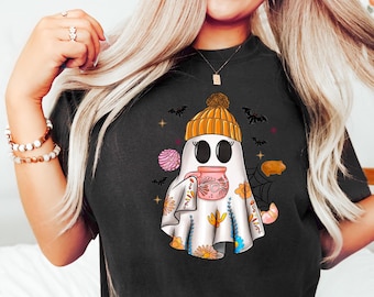 Könnte beinhalten: Schwarzes T-Shirt mit einem weißen Geist, der eine braune und orangefarbene Strickmütze trägt und eine rosa Tasse hält. Der Geist ist von Fledermäusen, Sternen und Süßigkeiten umgeben.