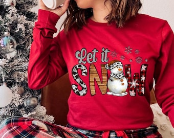 May include: Red long-sleeved shirt with a snowman graphic and the text "Let it Snow". The shirt has a plaid pattern and a leopard print design.
