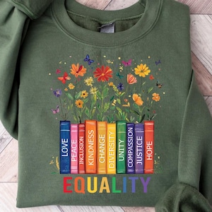 May include: Green sweatshirt with a rainbow stack of books that have words like "love", "peace", "inclusion", "change", "diversity", "unity", "compassion", "justice", and "hope". The words "EQUALITY" are printed below the books. The books are surrounded by colorful flowers.