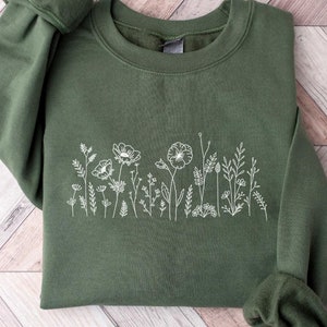 May include: A dark green sweatshirt with a white embroidered design of wildflowers. The design features a row of delicate flowers and leaves.