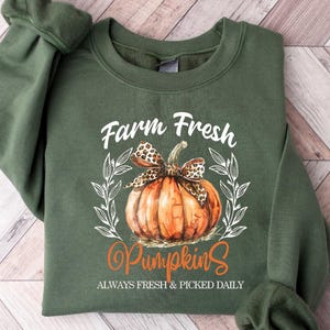 May include: Green sweatshirt with a graphic of an orange pumpkin with a leopard print bow, surrounded by leaves. The text reads "Farm Fresh Pumpkins" and "Always Fresh & Picked Daily" in white and orange.