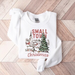 May include: White sweatshirt with a red and black graphic design that says "Small Town Christmas". The graphic features a church, houses, and a Christmas tree.