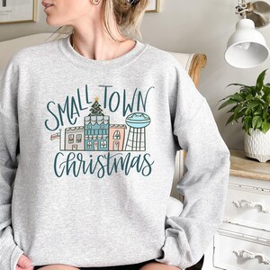 May include: A gray sweatshirt with the text "Small Town Christmas" in black and white lettering. The design features a small town with houses, a Christmas tree, and a water tower.