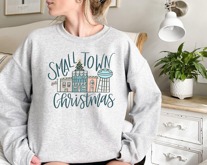 May include: A gray sweatshirt with the text "Small Town Christmas" in black and white lettering. The design features a small town with houses, a Christmas tree, and a water tower.