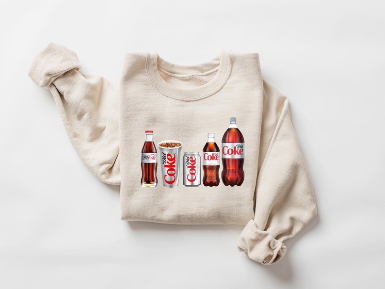 May include: A beige sweatshirt with a graphic print of four Coca-Cola products: a glass bottle of Coca-Cola, a can of Diet Coke, a can of Coca-Cola, and a glass bottle of Coca-Cola Zero Sugar.