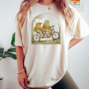 May include: A cream colored t-shirt with a graphic of two frogs riding a tandem bicycle. The frogs are wearing clothes and the bicycle is surrounded by green foliage.