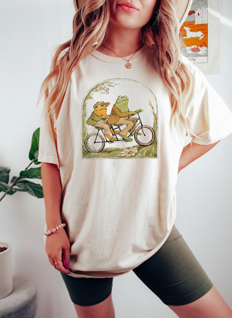 May include: A cream colored t-shirt with a graphic of two frogs riding a tandem bicycle. The frogs are wearing clothes and the bicycle is surrounded by green foliage.
