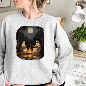May include: A grey sweatshirt featuring a graphic print of two ghosts sitting by a campfire in a forest. The moon is visible in the sky above the trees.