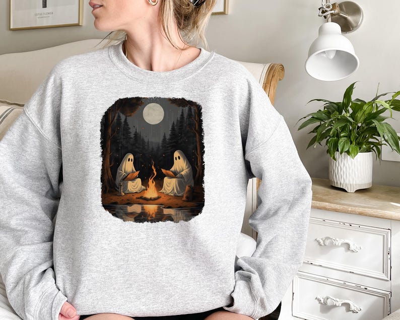 May include: A grey sweatshirt featuring a graphic print of two ghosts sitting by a campfire in a forest. The moon is visible in the sky above the trees.