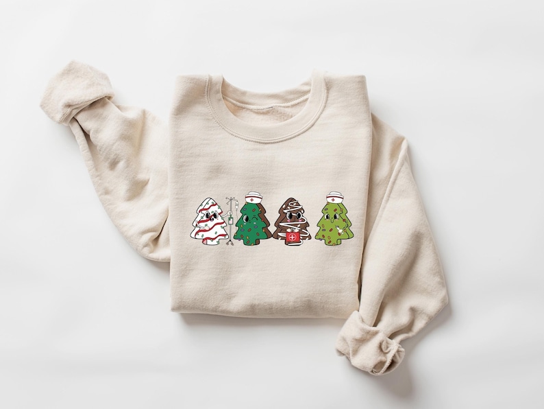 May include: A beige sweatshirt with a graphic of four Christmas trees dressed as nurses. The trees are wearing red and white striped hats, green hats, and brown hats. One tree is holding a red cross.