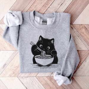 May include: A gray sweatshirt with a black cat eating ramen noodles. The cat is holding chopsticks and has a bowl of ramen in front of it.