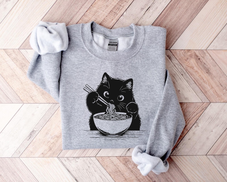 May include: A gray sweatshirt with a black cat eating ramen noodles. The cat is holding chopsticks and has a bowl of ramen in front of it.