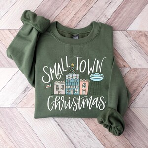 May include: A green sweatshirt with the text "Small Town Christmas" in white lettering. The design features a small town with houses, a Christmas tree, and a star.