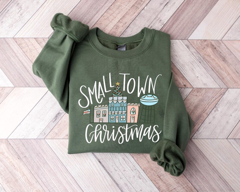 May include: A green sweatshirt with the text "Small Town Christmas" in white lettering. The design features a small town with houses, a Christmas tree, and a star.