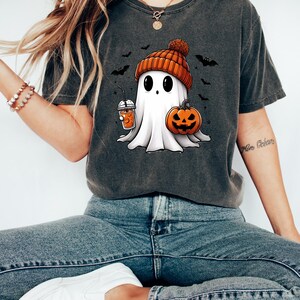 May include: A dark gray t-shirt with a cartoon ghost wearing an orange and brown knit hat holding a pumpkin and a cup of orange juice. The ghost is surrounded by bats.