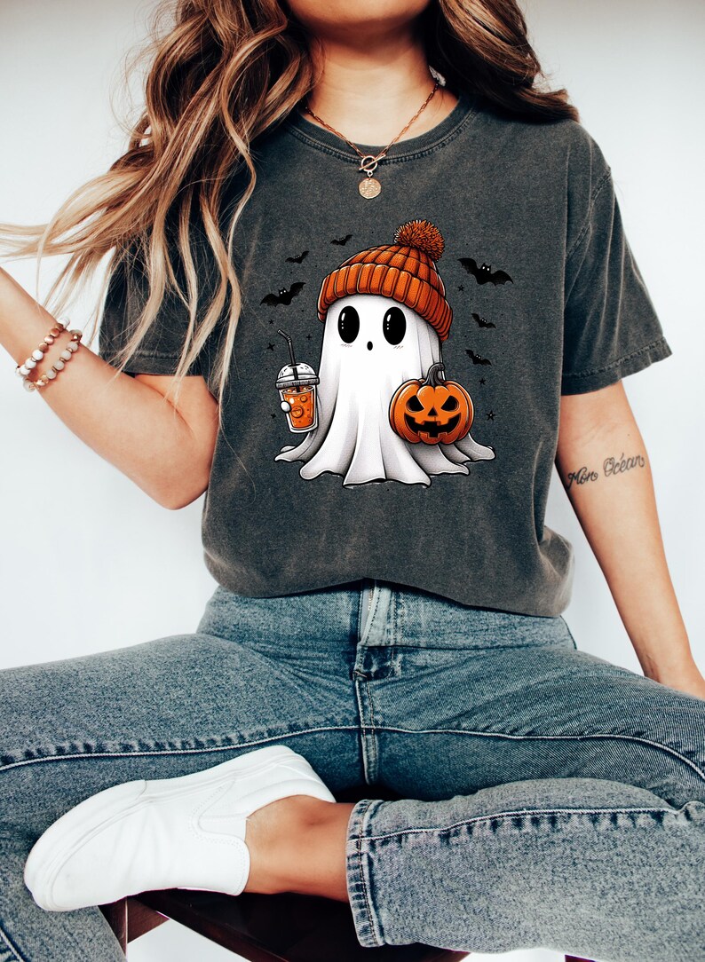 May include: A dark gray t-shirt with a cartoon ghost wearing an orange and brown knit hat holding a pumpkin and a cup of orange juice. The ghost is surrounded by bats.