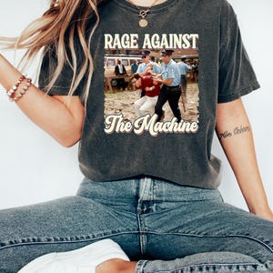 K&ouml;nnte beinhalten: Ein dunkelgraues T-Shirt mit einer Grafik im Vintage-Stil der Band Rage Against The Machine. Die Grafik zeigt ein Schwarzwei&szlig;foto eines Mannes, der von zwei Polizisten verhaftet wird. Der Text "Rage Against The Machine" ist in Gelb &uuml;ber dem Foto gedruckt, und der Name der Band ist in einer Retro-Schrift unter dem Foto gedruckt.