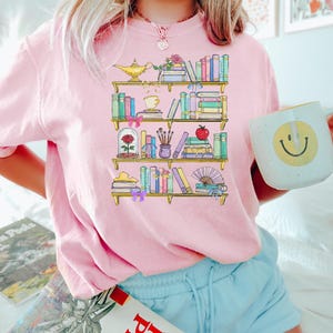 May include: A pink t-shirt with a graphic of a bookshelf filled with books, a magic lamp, a rose, an apple, and other decorative items. The shirt is being worn by a person with blonde hair and blue shorts.