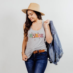 Puede incluir: Un top gris sin mangas con la palabra "NURSE" en letras de colores y un dise&ntilde;o floral. El top lo lleva una persona con el pelo largo y casta&ntilde;o y un sombrero de paja. La persona tambi&eacute;n lleva pantalones vaqueros azules y una chaqueta vaquera.