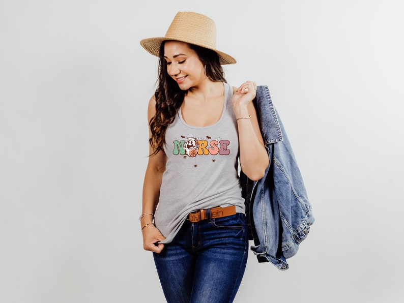 Puede incluir: Un top gris sin mangas con la palabra "NURSE" en letras de colores y un dise&ntilde;o floral. El top lo lleva una persona con el pelo largo y casta&ntilde;o y un sombrero de paja. La persona tambi&eacute;n lleva pantalones vaqueros azules y una chaqueta vaquera.