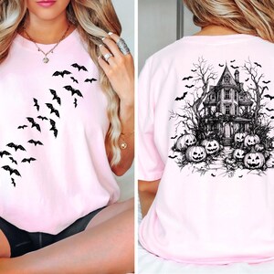 Op de afbeelding: Roze T-shirt met een zwart vleermuisontwerp aan de voorkant en een zwart spookhuis- en pompoenontwerp aan de achterkant.