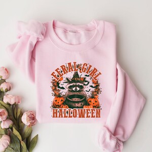 Pu&ograve; includere: Felpa rosa con una grafica di un procione che indossa un cappello da strega e tiene un calderone. Il testo "Feral Girl Halloween" &egrave; stampato sotto la grafica.