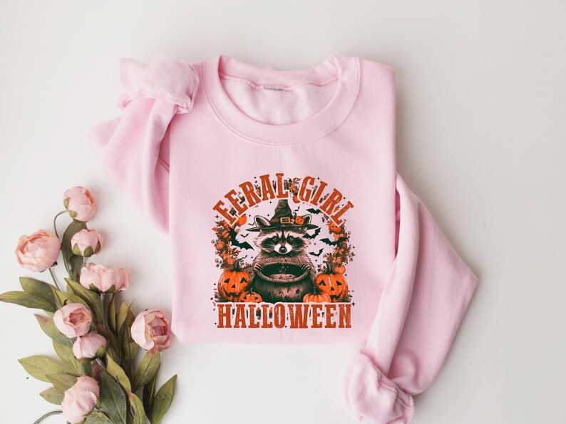 Pu&ograve; includere: Felpa rosa con una grafica di un procione che indossa un cappello da strega e tiene un calderone. Il testo "Feral Girl Halloween" &egrave; stampato sotto la grafica.