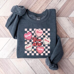 May include: A dark gray sweatshirt with a graphic of pink and white hearts on a checkered background. The hearts have words on them, including "You are", "Loved", "Enough", "Strong", "Capable", and "Worthy".
