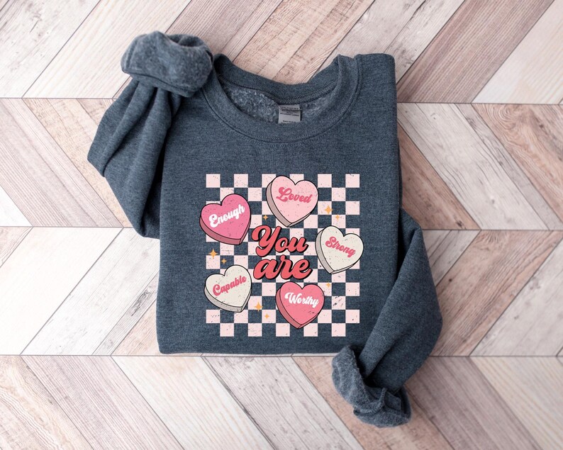 May include: A dark gray sweatshirt with a graphic of pink and white hearts on a checkered background. The hearts have words on them, including "You are", "Loved", "Enough", "Strong", "Capable", and "Worthy".