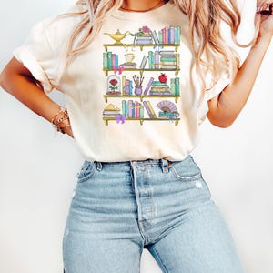 May include: A cream colored t-shirt with a graphic of a bookshelf filled with books, a rose, a lamp, and other decorative items.