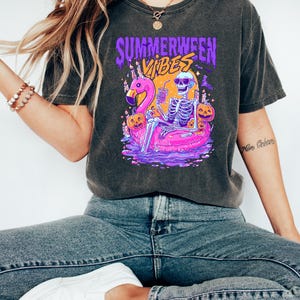 May include: Dark gray t-shirt with a graphic featuring a skeleton relaxing on a pink flamingo floatie, surrounded by pumpkins. The shirt has the text "Summerween Vibes" in purple and orange.